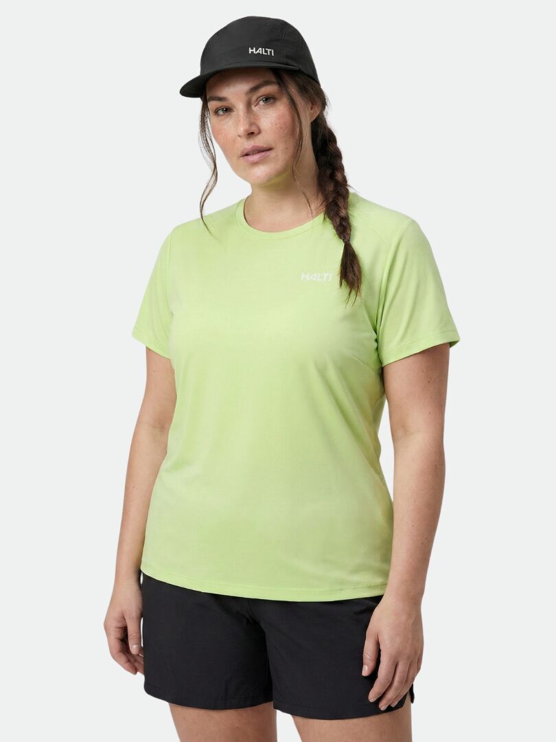 En person i ljusgrön Halti Salves T-skjorta Dam Plus, svarta shorts och svart keps står mot en enfärgad bakgrund.