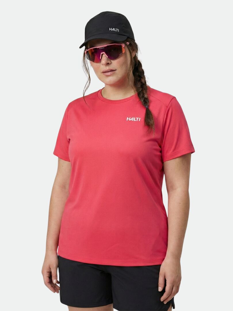 En kvinna i rosa Halti Salves T-skjorta Dam Plus, svarta shorts, keps och rosa solglasögon står mot en enfärgad bakgrund.