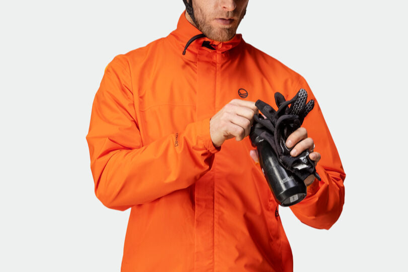 Person i orange jacka håller i en svart vattenflaska och ett par svarta handskar mot en enfärgad bakgrund.