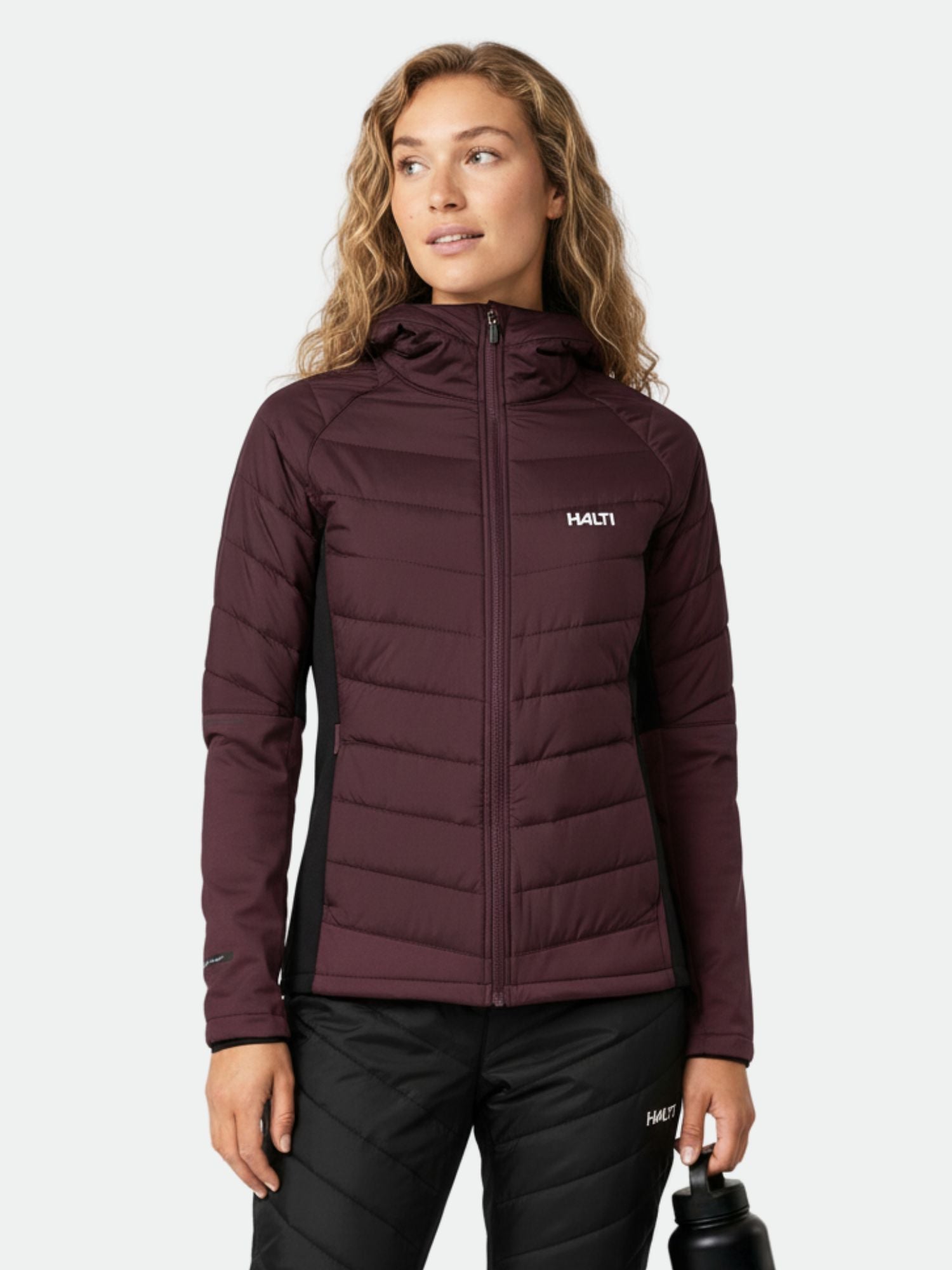 Person i rödbrun Halti Hanki 2 Hybrid Längdskidjacka Dam och svarta byxor, håller i en vattenflaska mot en ljus bakgrund.