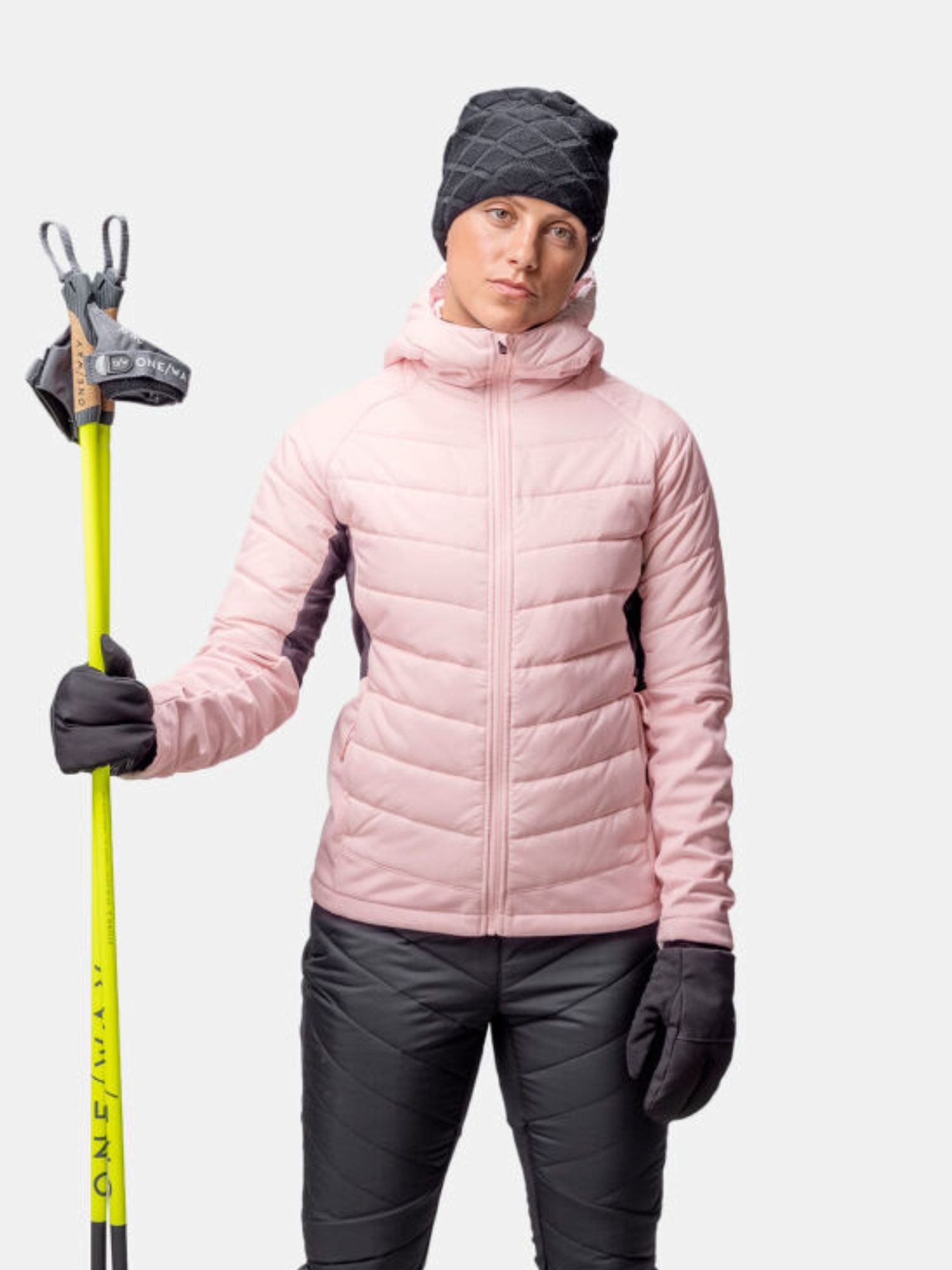 En person i vintersportkläder bär en rosa Halti Hanki 2 Hybrid Längdskidjacka Dam och håller i gula skidstavar.