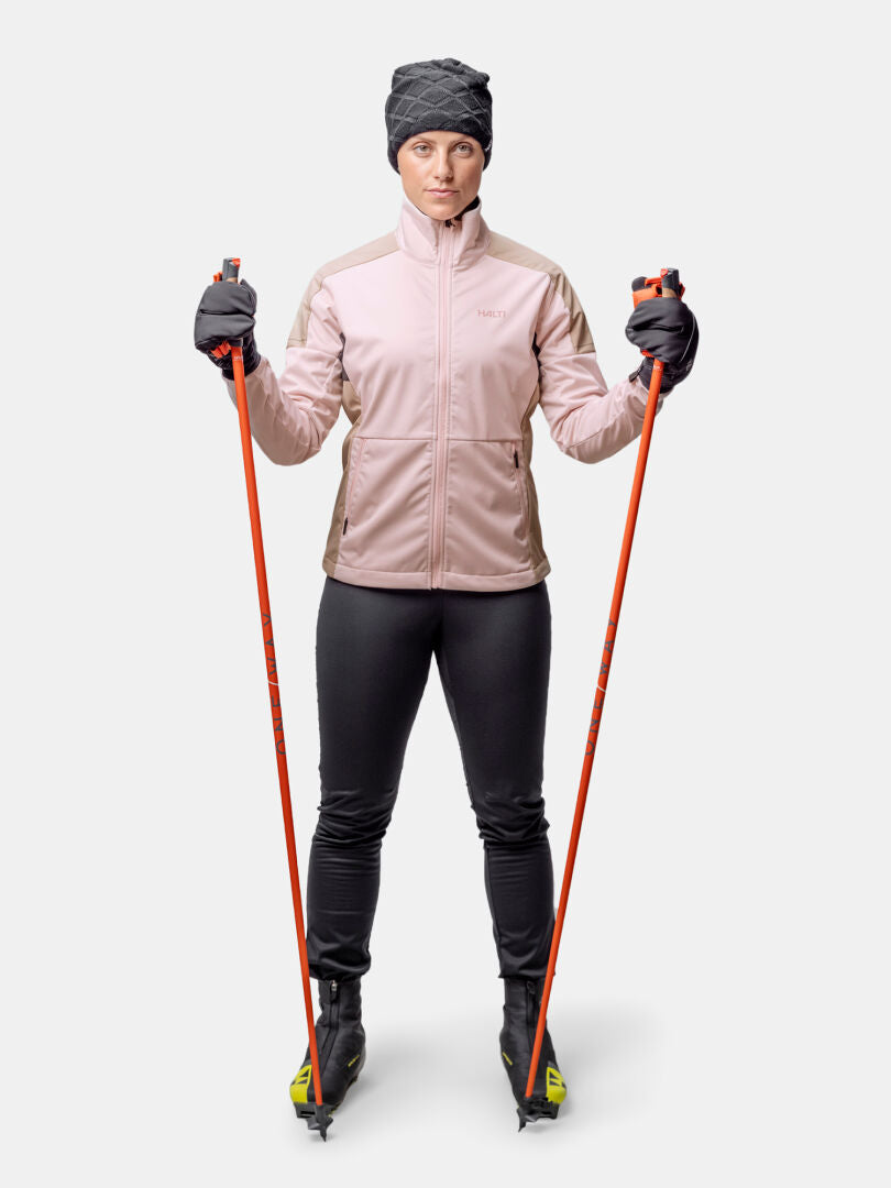 En person bär Halti Huuru Längdskiddräkt Dam, håller i orangefärgade skidstavar, med skidor, handskar, mössa och jacka.