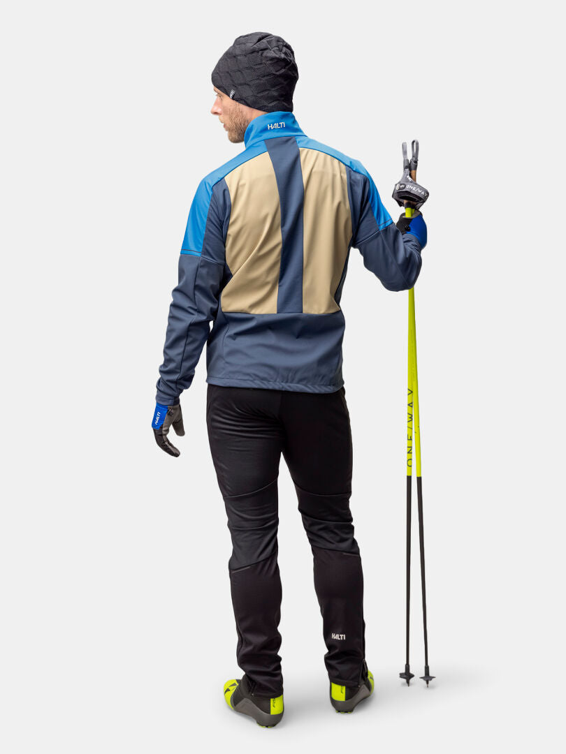 En man i Halti Framover Längdskidjacka Herr håller i skidstavar, vänd bortåt, och gör ett fredstecken med höger hand.