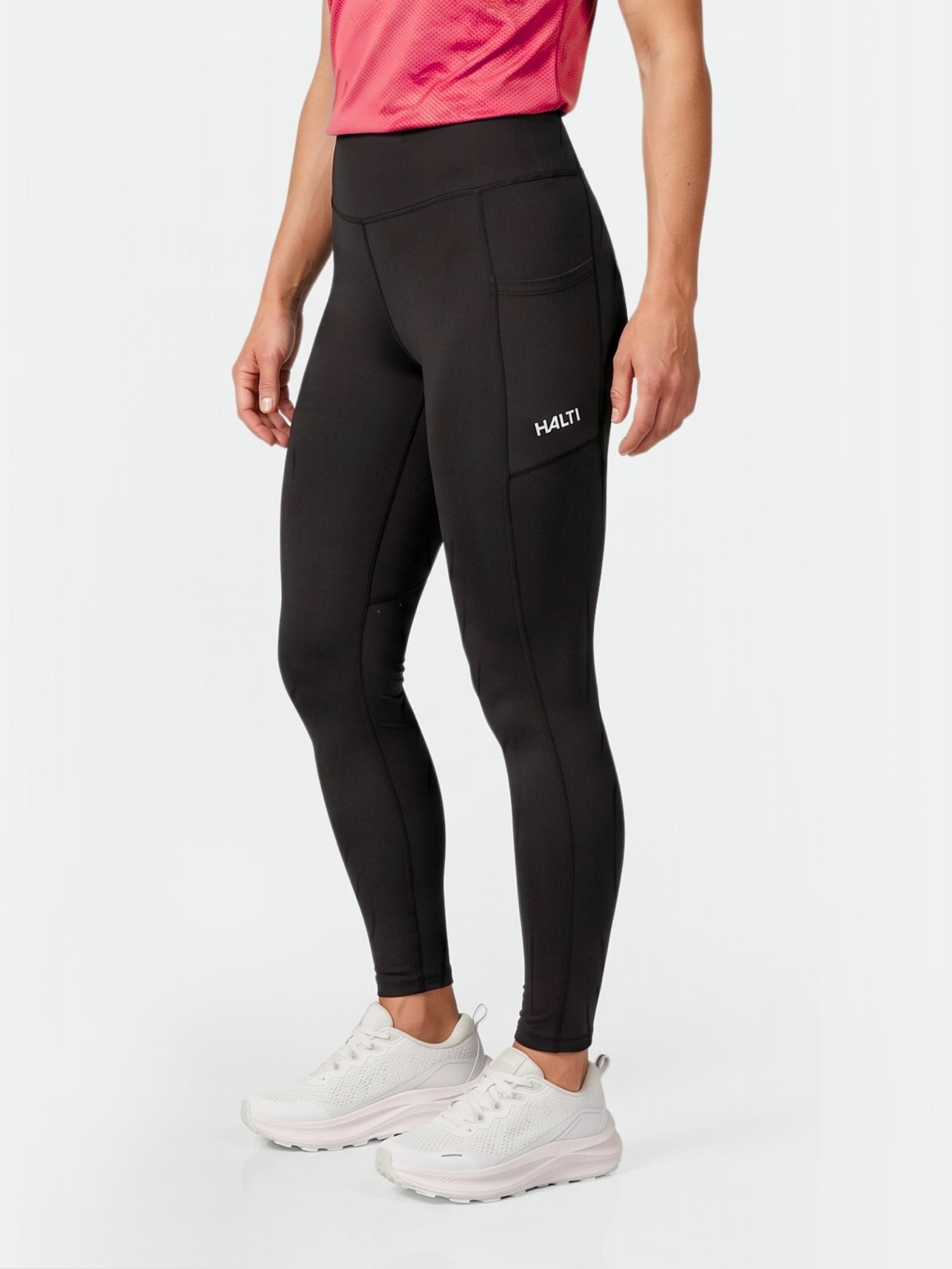 En person bär Halti Kaldu Träningstights Dam med fickor, rosa topp och vita sneakers mot en enkel bakgrund.