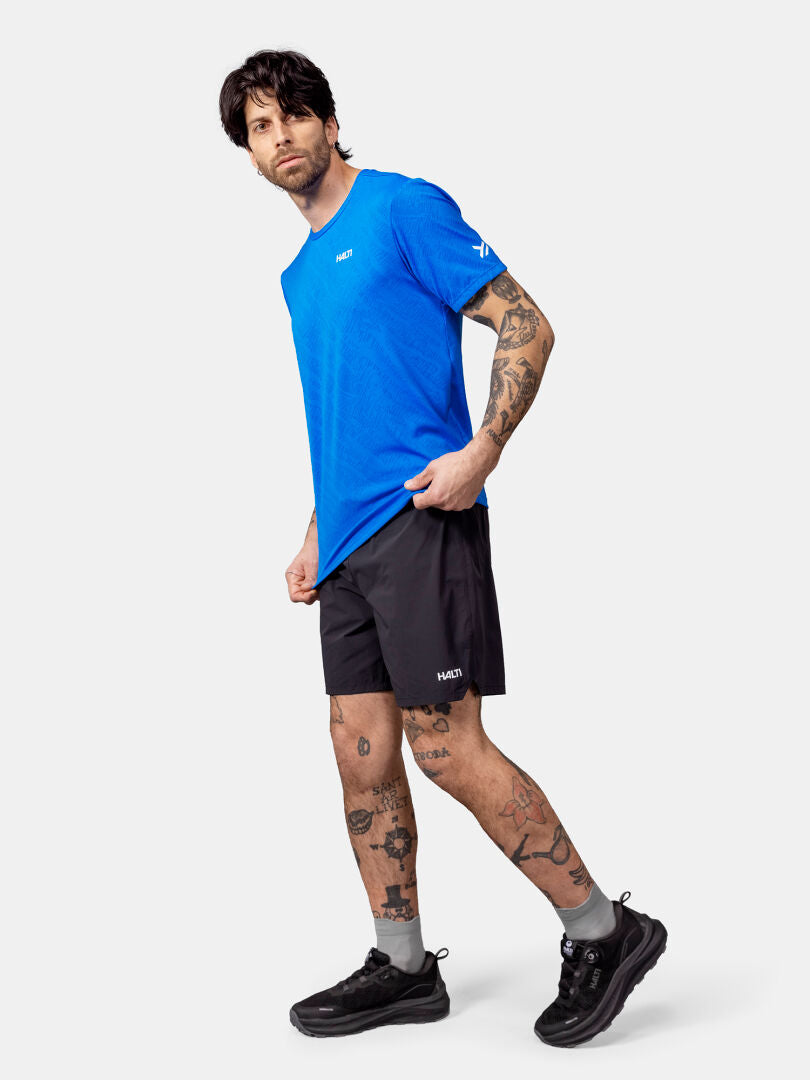 En tatuerad man bär en blå Halti Oulanka T-skjorta Herr, svarta shorts och sneakers mot en enfärgad bakgrund.