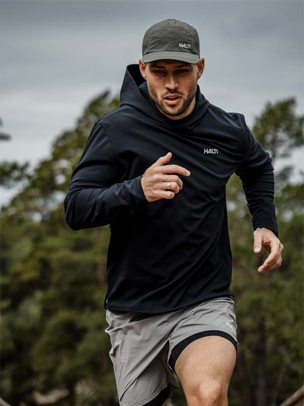 Man i Halti Kaltio Träningshoodie Herr och grå shorts springer ute, grön keps och träd i bakgrunden.