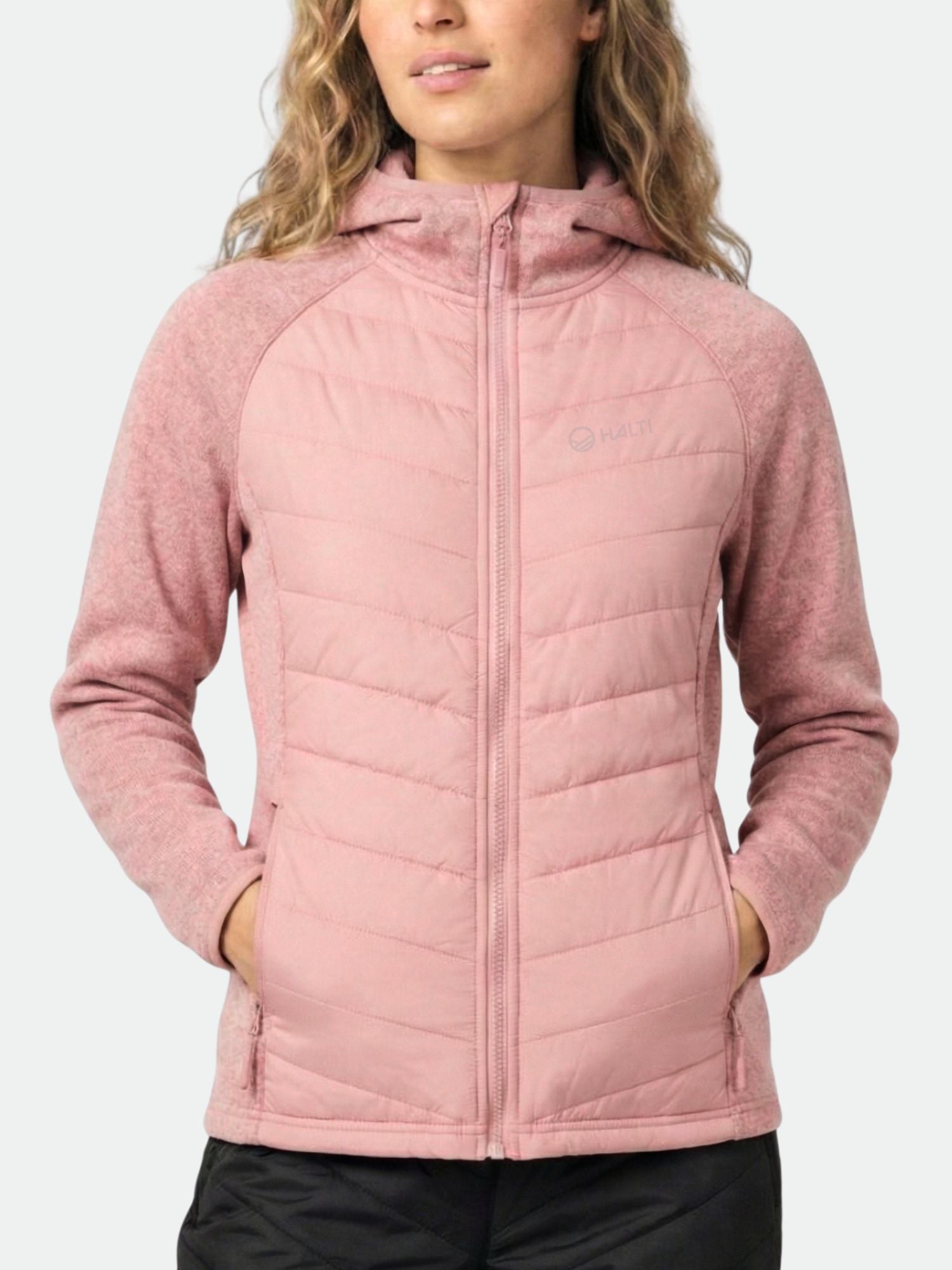 En kvinna bär Halti Streams Hybrid Mellanlager Dam i ljusrosa fleece med händerna i fickorna, midjan upp.