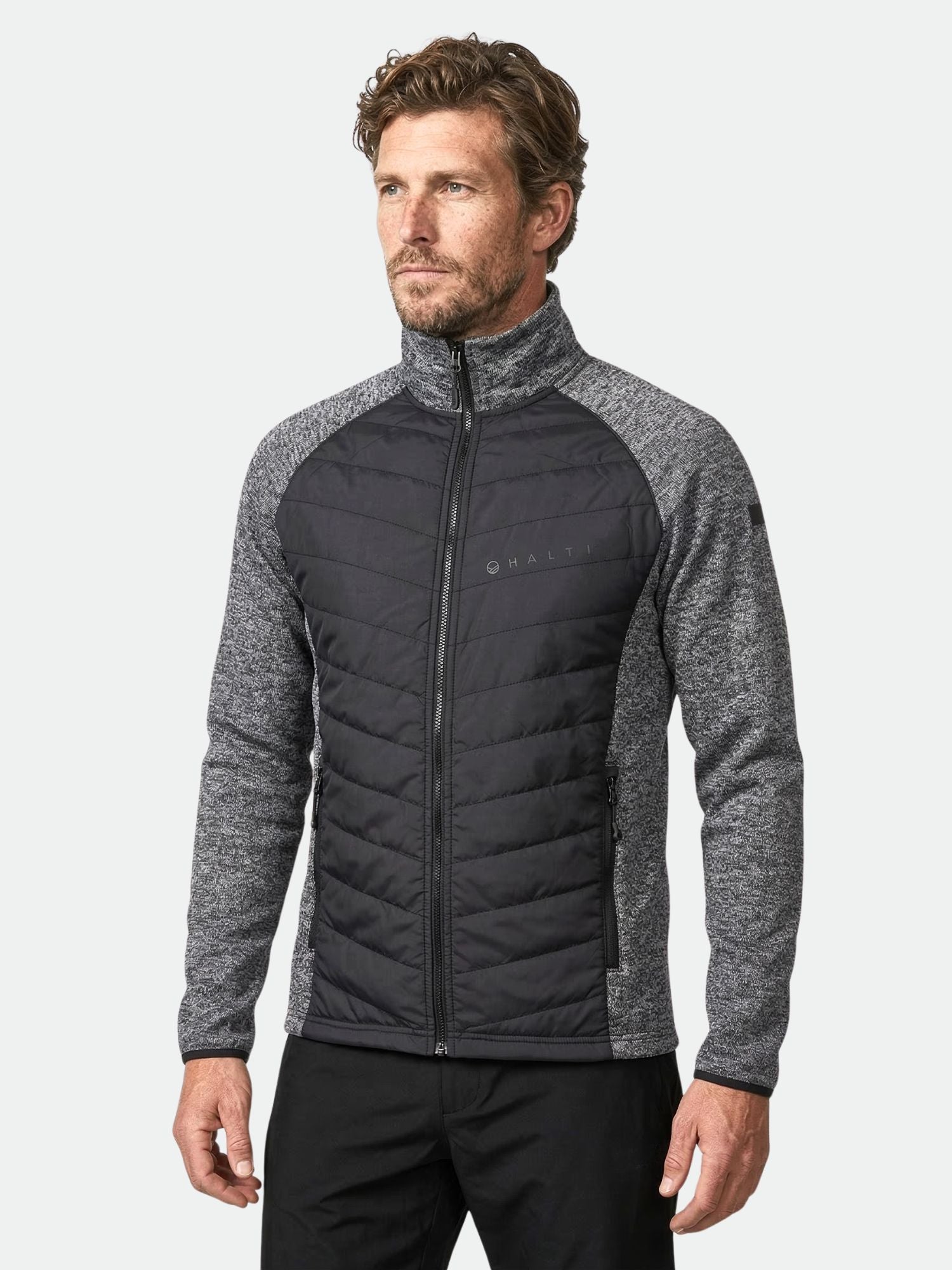 Man klädd i Halti Streams Hybrid Mellanlager Herr-jacka i grått och svart, med hög krage, mot ljusgrå bakgrund.