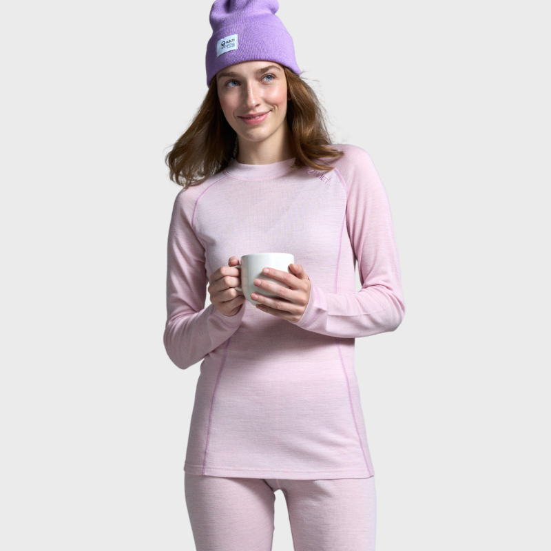 Kvinna i ljusrosa Halti Hossa Merinoull Underställ Set och en lila mössa håller en mugg mot en enfärgad bakgrund.