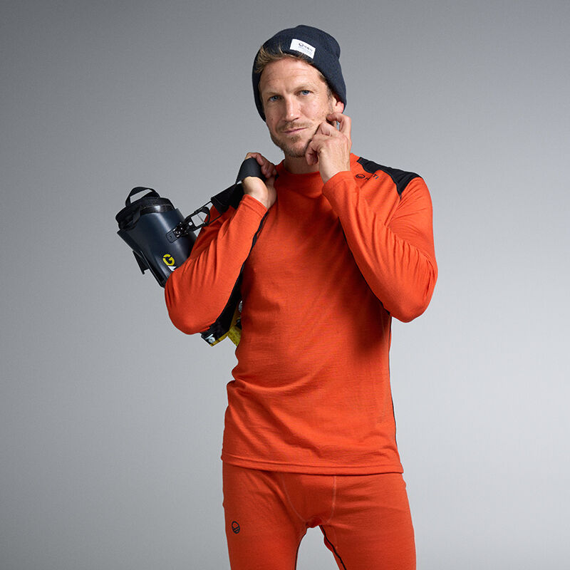 Man i orange underställsdräkt håller pjäxor över axeln och står mot en grå bakgrund.