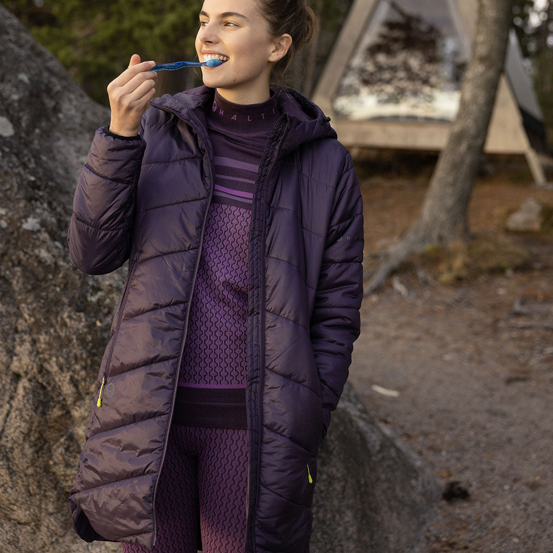 En person i en lila Halti Element Parka Dam borstar tänderna utomhus nära ett tält och träd.