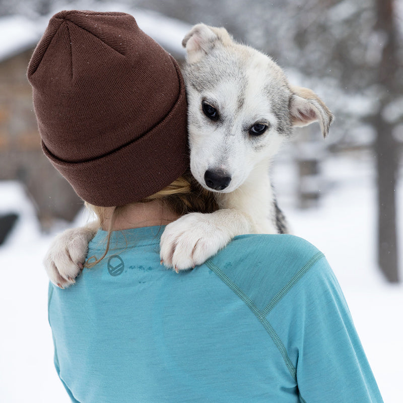 En kvinna i blå skjorta och brun mössa håller en hund, iklädd Halti Pihka II Merinoull Underställströja Dam utomhus.