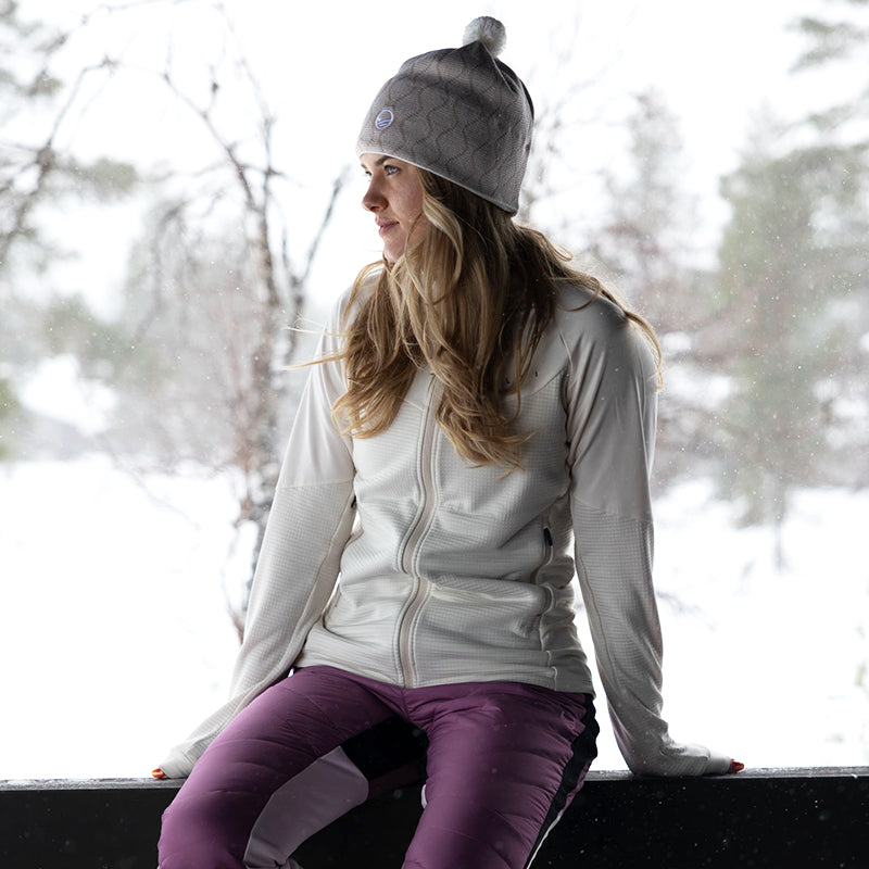 En kvinna i en Halti HeatGrid Mellanlager Dam och lila byxor sitter på en snöig bänk, med träd bakom sig.