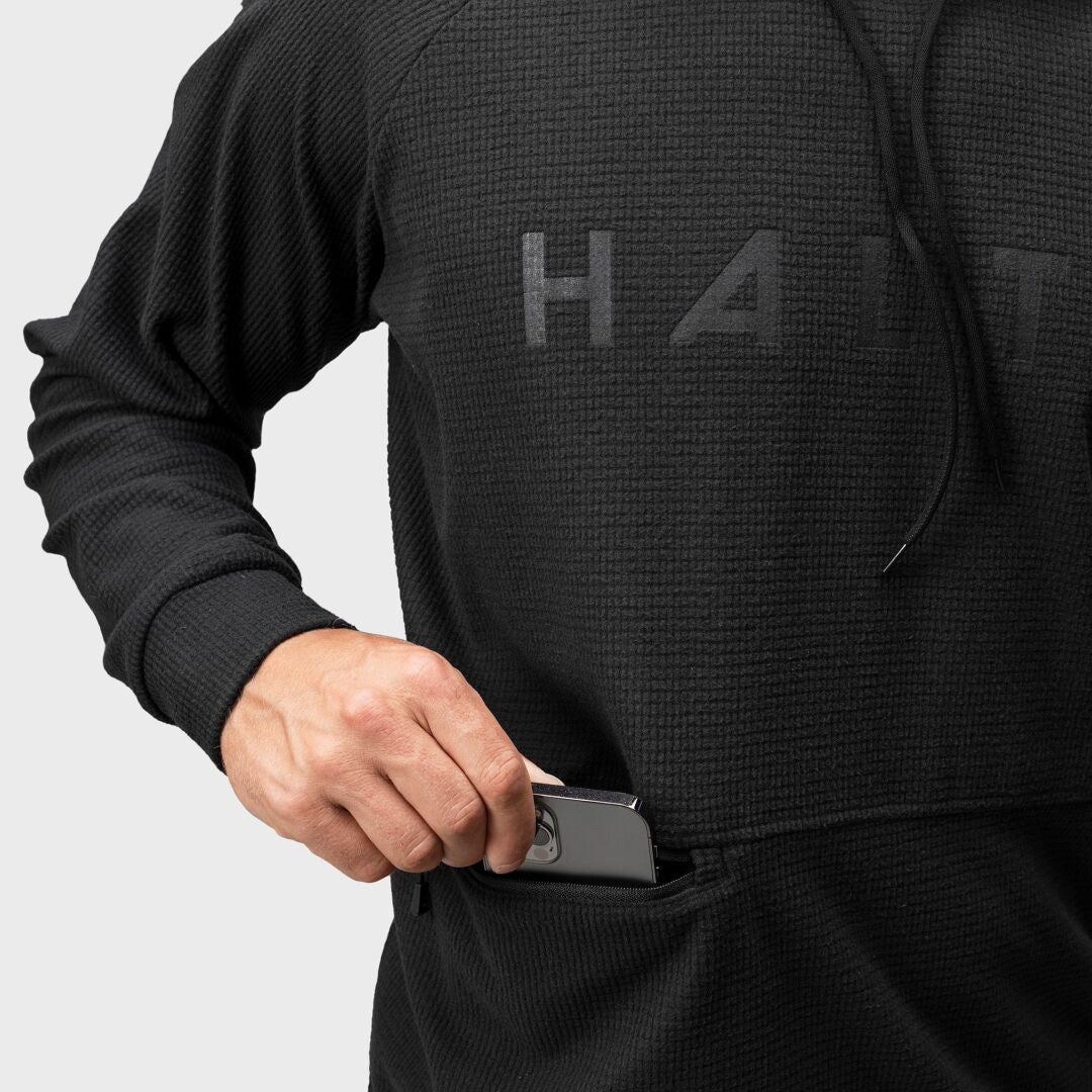 En person i en Halti Crissgon Hoodie Herr tillverkad av återvunnen polyester stoppar en smartphone i framfickan.