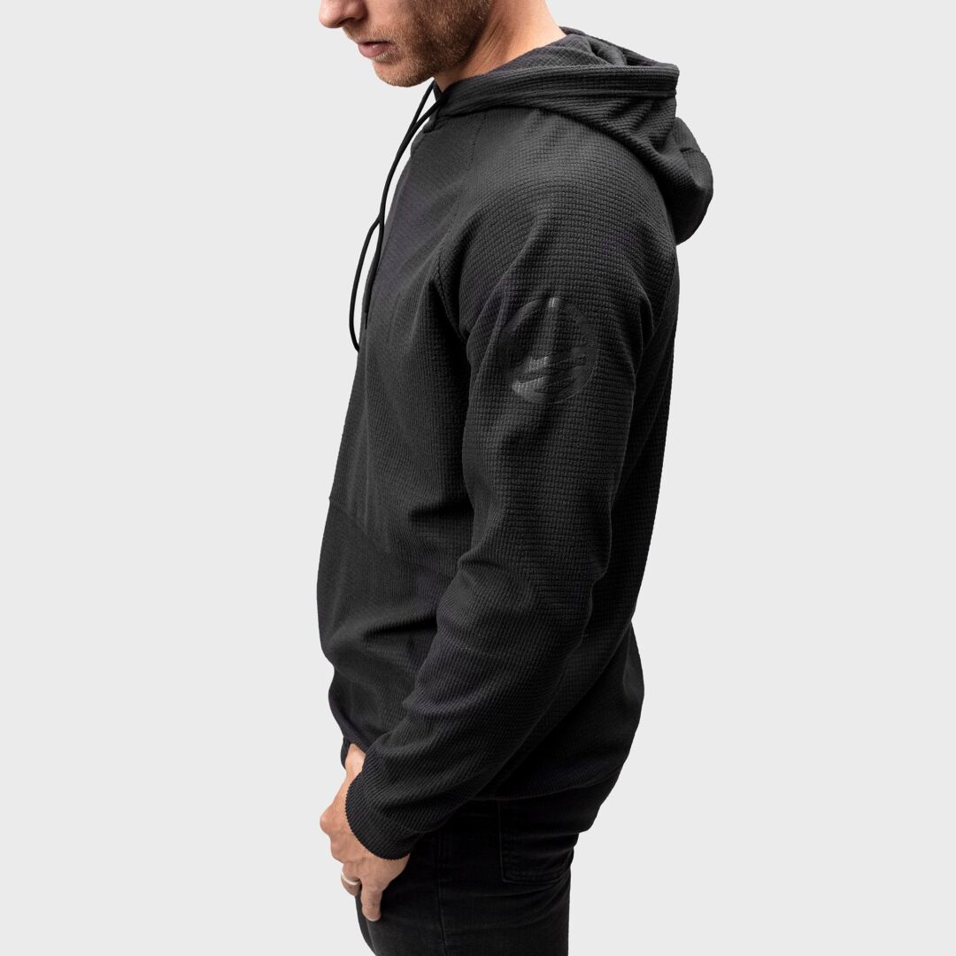 En man i en svart Crissgon Hoodie Herr från Halti, tillverkad med Active Dry-tyg, står snett mot en ljusgrå bakgrund.