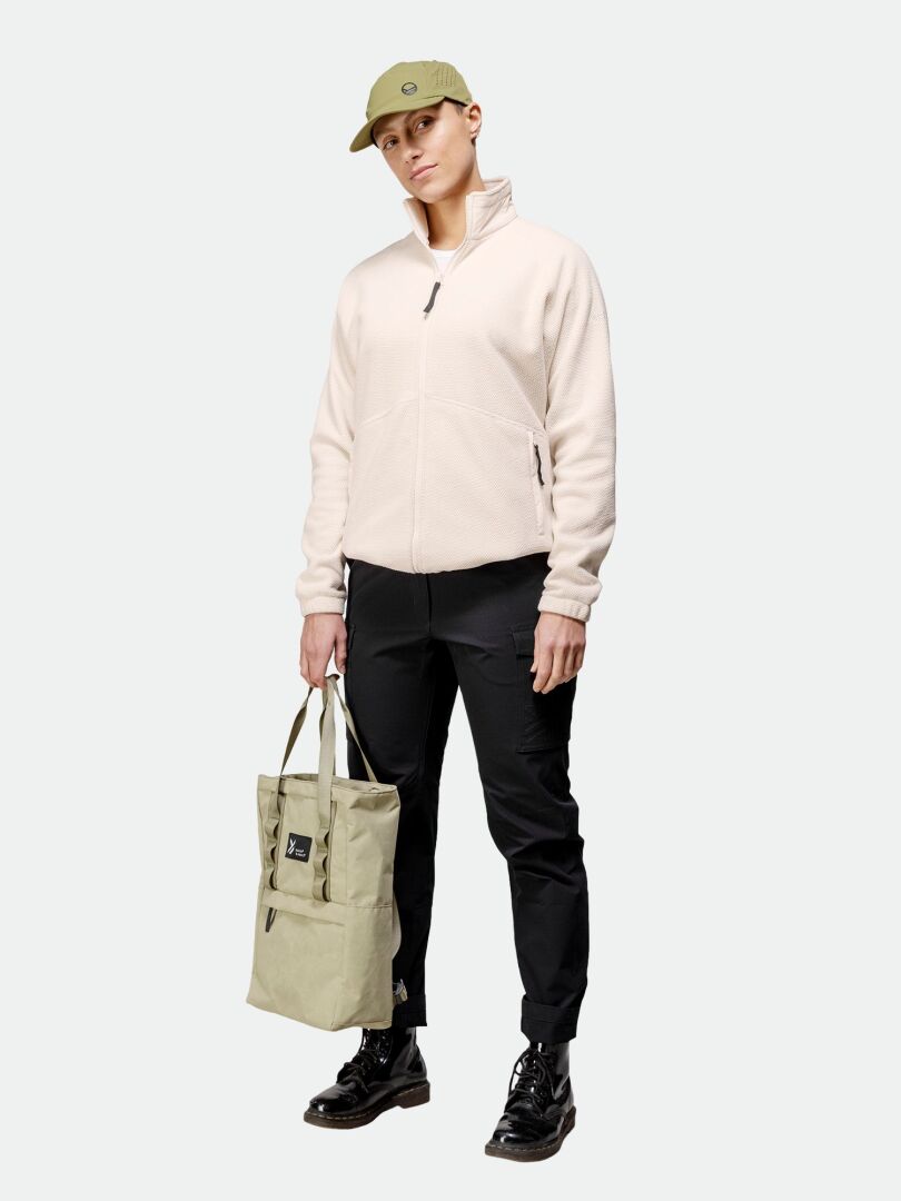 En person klädd i beige Halti Soolo Mellanlager Dam-jacka, svarta byxor, grön keps och svarta skor håller i en beige tygväska.