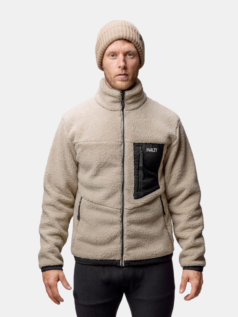 Man i Halti Lumista Polar Fleecejacka med svarta dragkedjor och beige mössa mot enkel bakgrund.