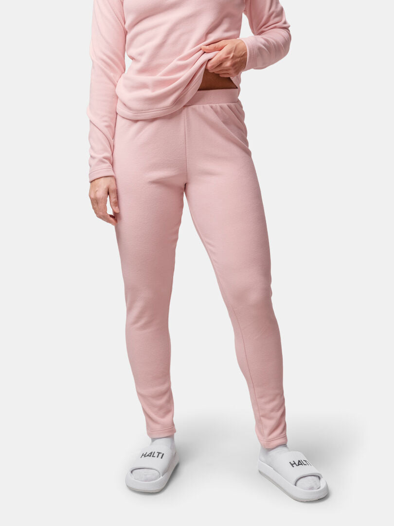 En kvinna bär Halti Trone Fleece Byxor Dam i ljusrosa, står mot en vit bakgrund och lyfter skjortan något.