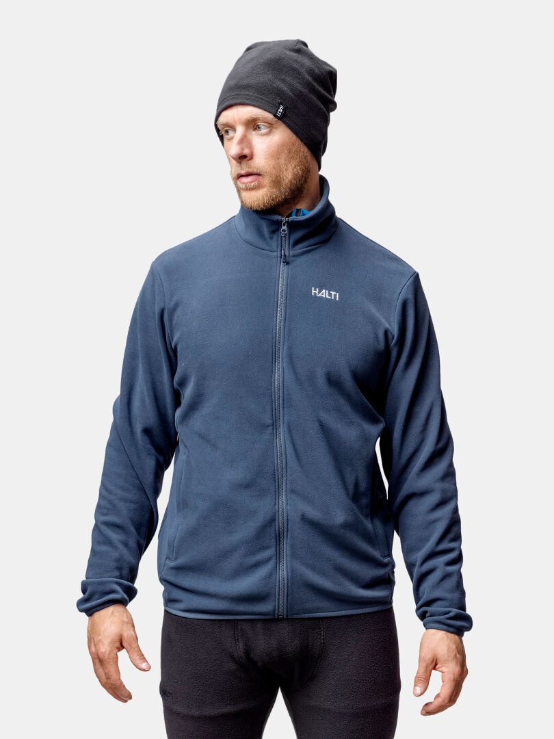 En man i mörk mössa och Halti Rockmoon Fleecejacka Herr står mot en vanlig ljus bakgrund.