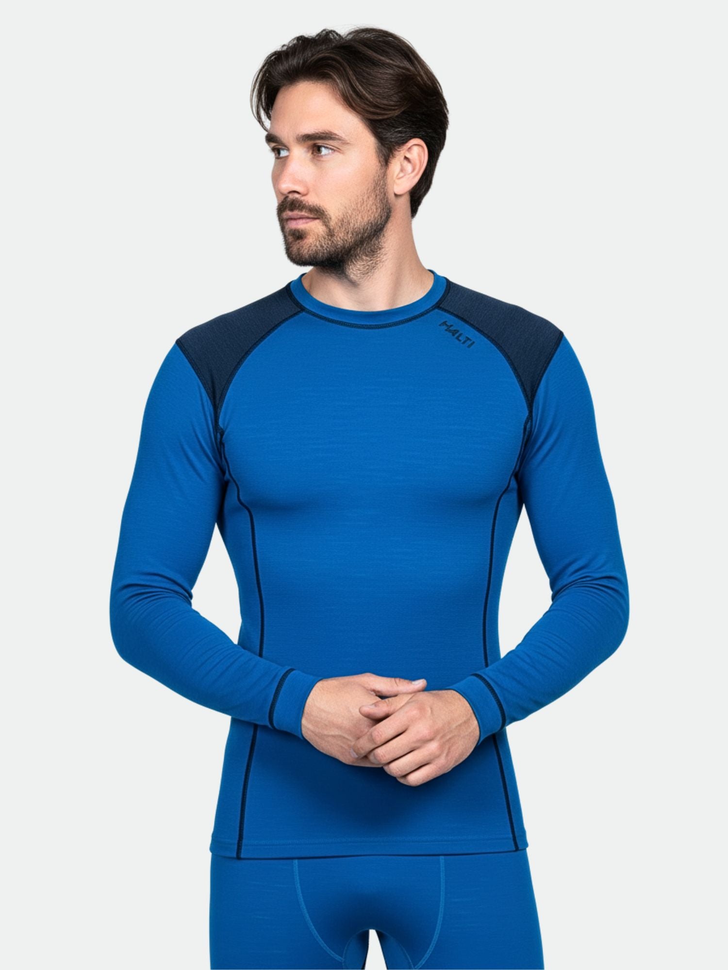 En man bär Halti Hossa II Merino Underställ Set Herr, som består av en blå topp med svarta axlar och matchande byxor.