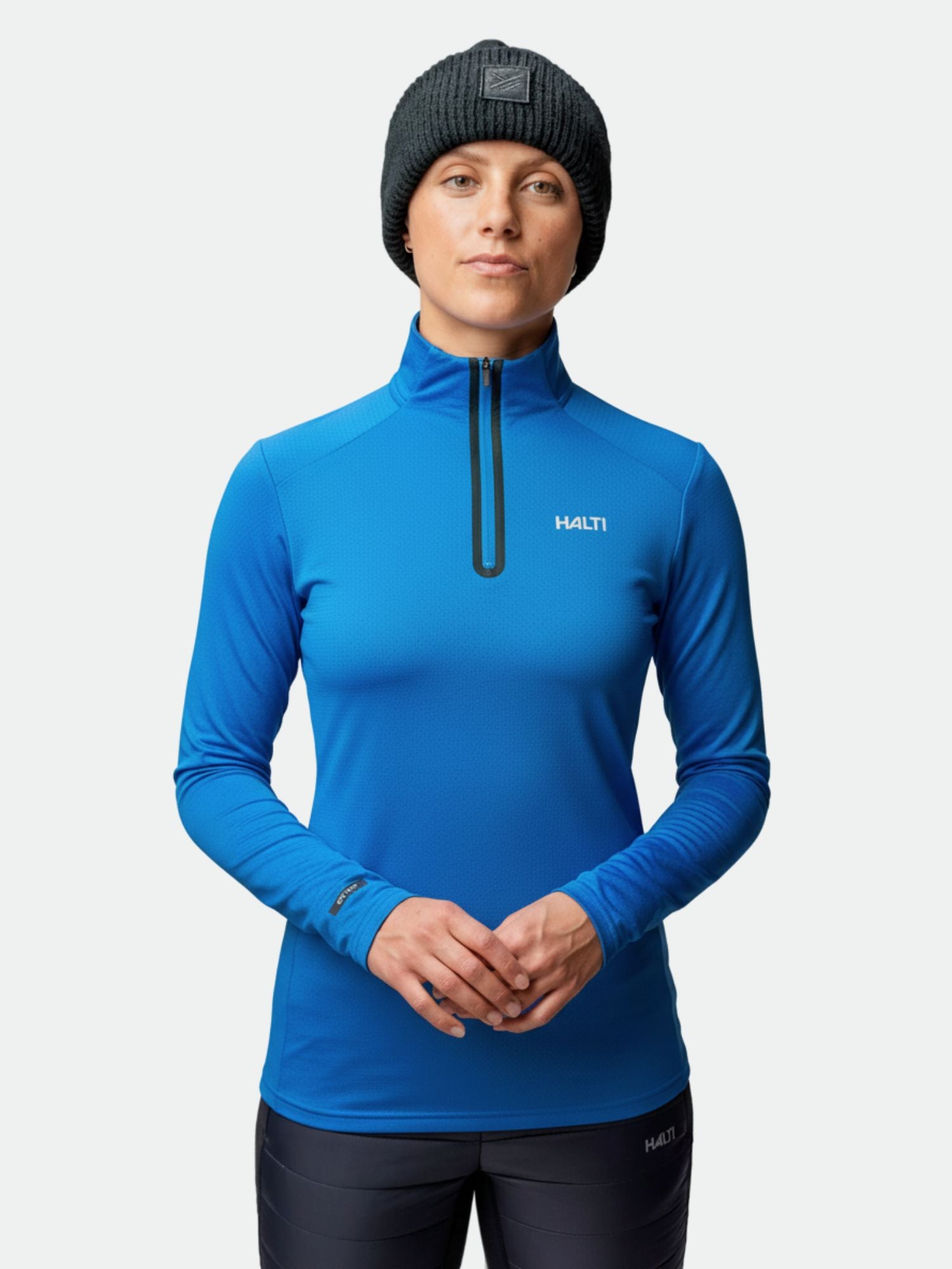 En person i blå Halti Jaako Tröja Dam-topp och svart mössa vänder sig mot kameran mot en enfärgad bakgrund.