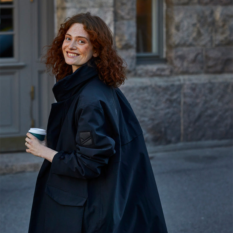 En person med lockigt rött hår och mörk rock håller i en to-go-mugg och ler när hon går på en gata i staden.