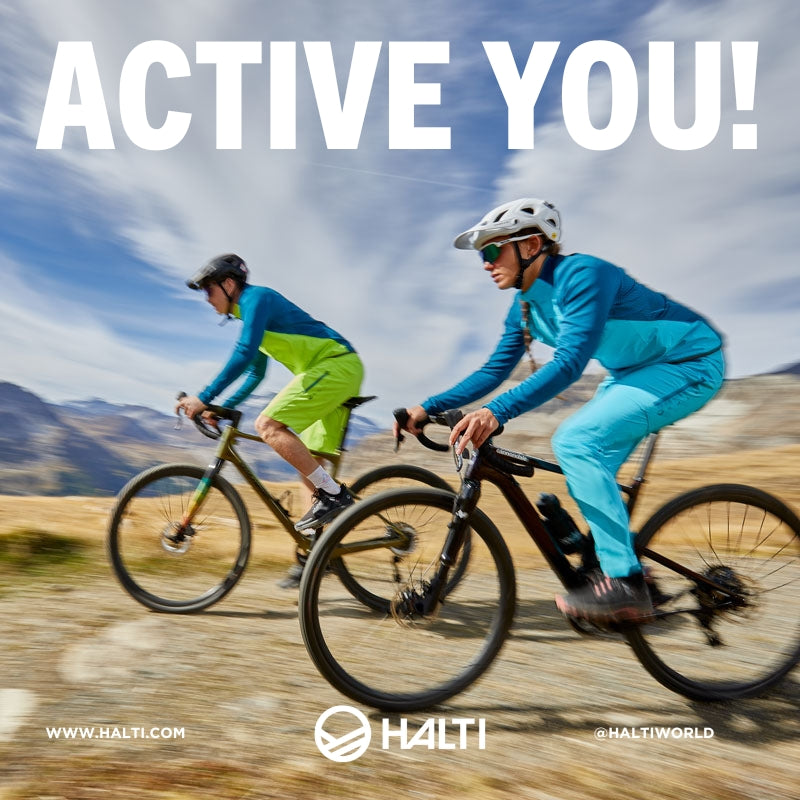 Två personer i hjälm och sportkläder cyklar mountainbike på en stig med berg och blå himmel i bakgrunden.