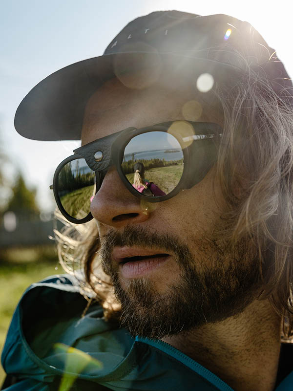 Närbild på en man med solglasögon och keps utomhus, med en kvinna som speglar sig i hans solglasögon.