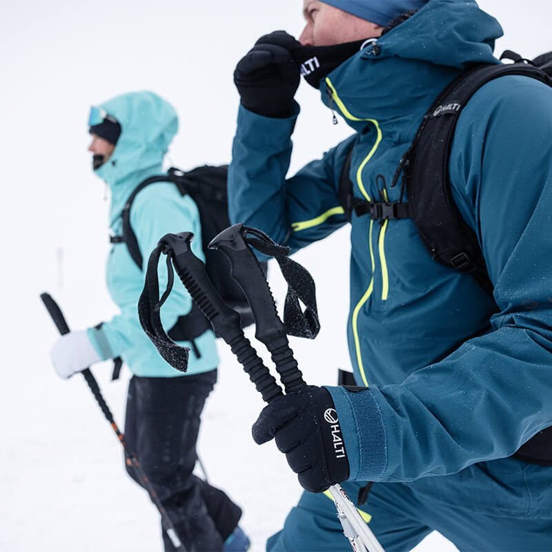 Två personer i vinterkläder med skidstavar går genom snöig terräng; snön faller och sikten är låg.