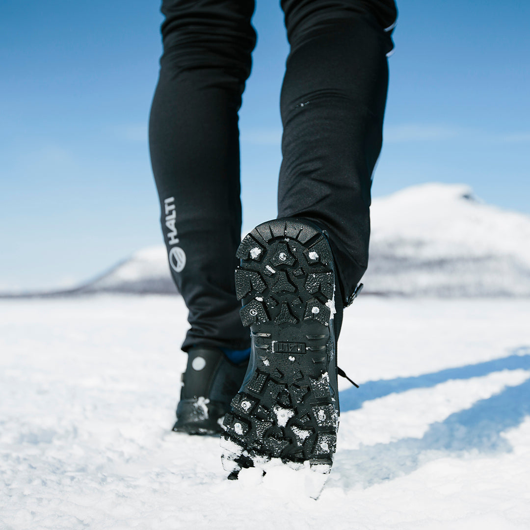 Närbild av en person som går på snö i dubbade stövlar, med berg och blå himmel i bakgrunden.
