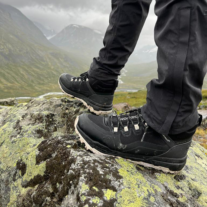 Person i svarta vandringskängor och byxor står på en mossig klippa med berg och dalar i bakgrunden.