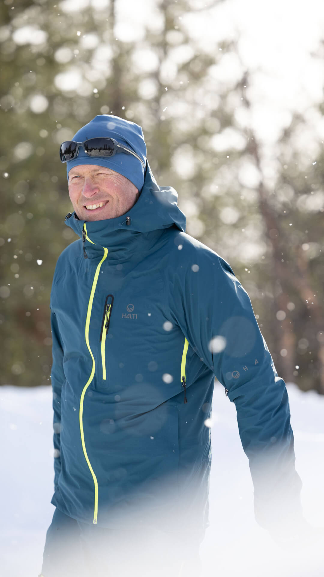 En person i blå vinterjacka och solglasögon står utomhus i ett snöigt landskap med träd i bakgrunden.
