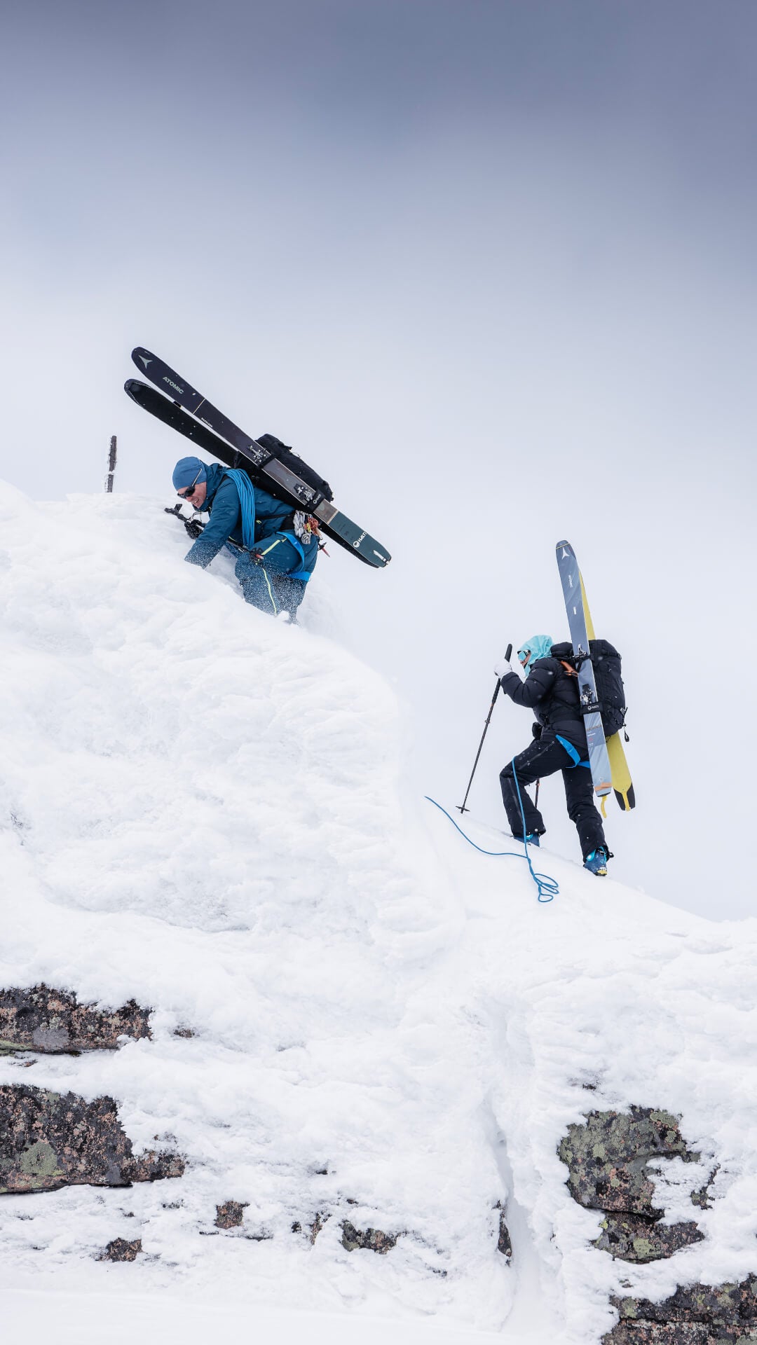Två personer i vinterkläder klättrar uppför en snöig sluttning med skidor på ryggsäckarna under en molnig himmel.