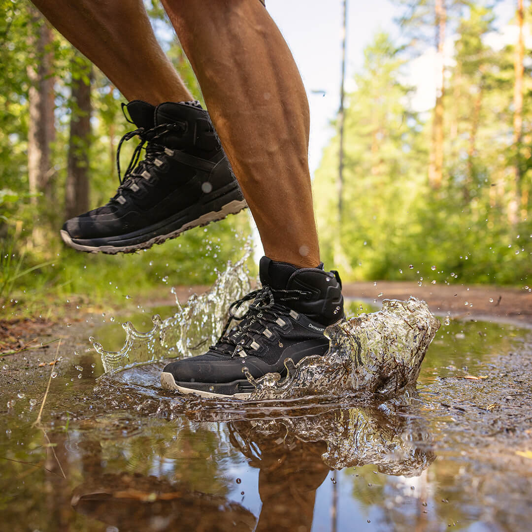 En person i svarta vandringskängor hoppar ner i en lerpöl på en skogsstig och sprutar vatten omkring sig.