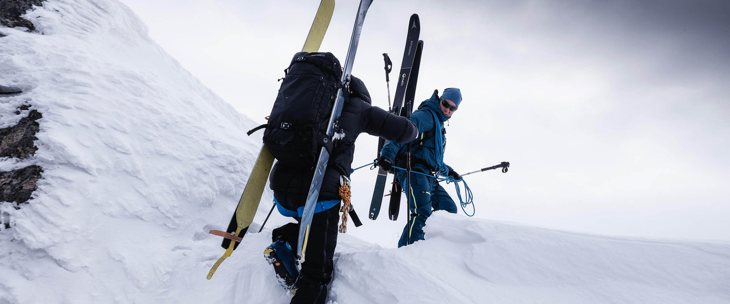 Två personer i vinterkläder bär skidor och stavar medan de klättrar uppför en snöig bergssluttning under en molnig himmel.
