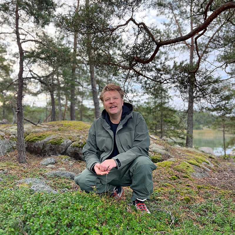 En person i gröna friluftskläder står på knä på mossig mark i ett skogsområde med träd och stenar.