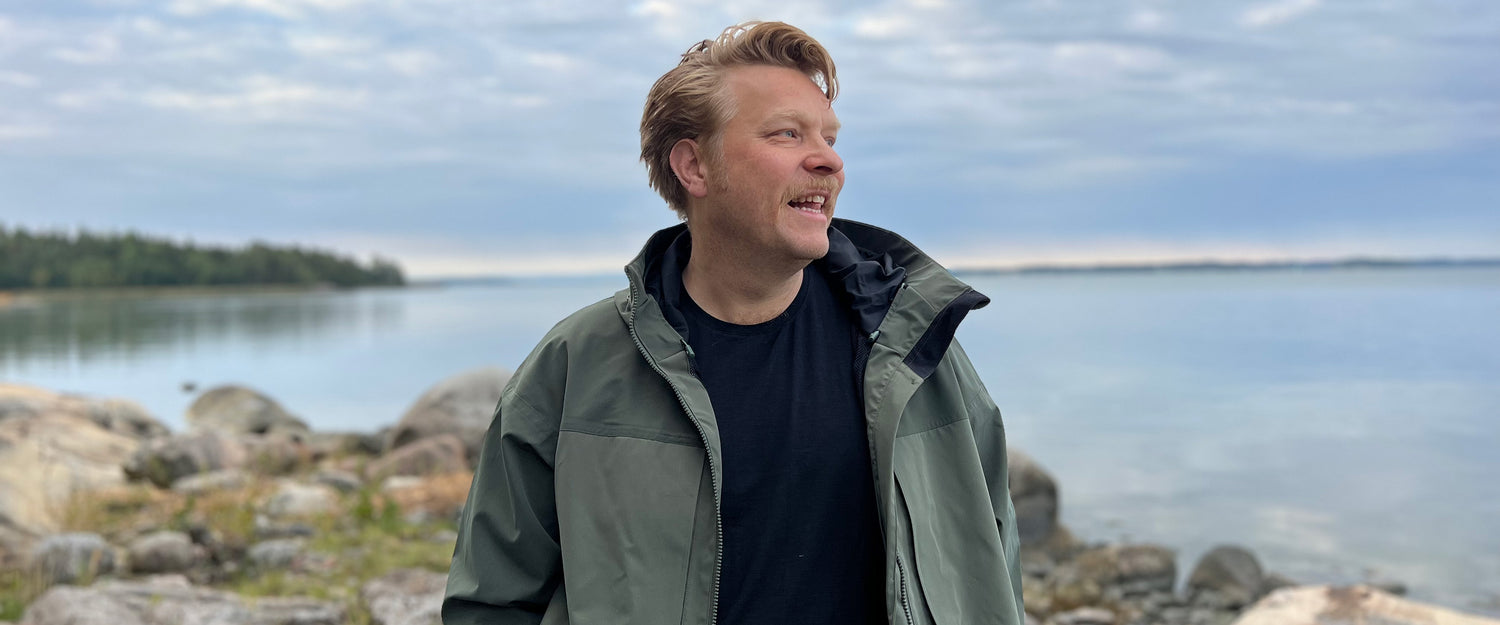 En man i grön jacka står vid en klippig strand med lugnt vatten och molnig himmel i bakgrunden.