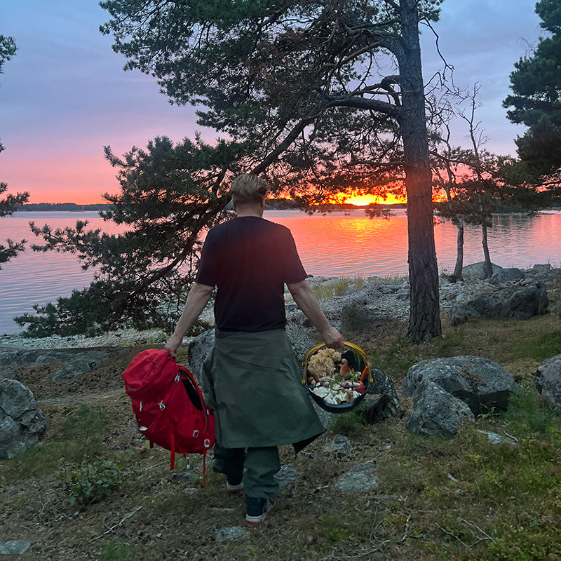 En person går vid en sjö i solnedgången, med en röd ryggsäck och en korg med svamp, omgiven av träd och stenar.