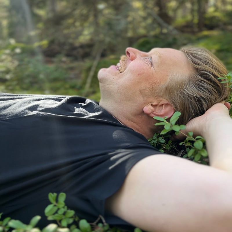 En man ligger på rygg utomhus i en skog, tittar upp och ler med ena handen bakom huvudet.