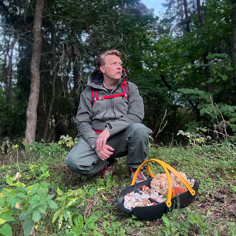 En person i friluftskläder står på knä i en skog bredvid en korg fylld med olika svampar.