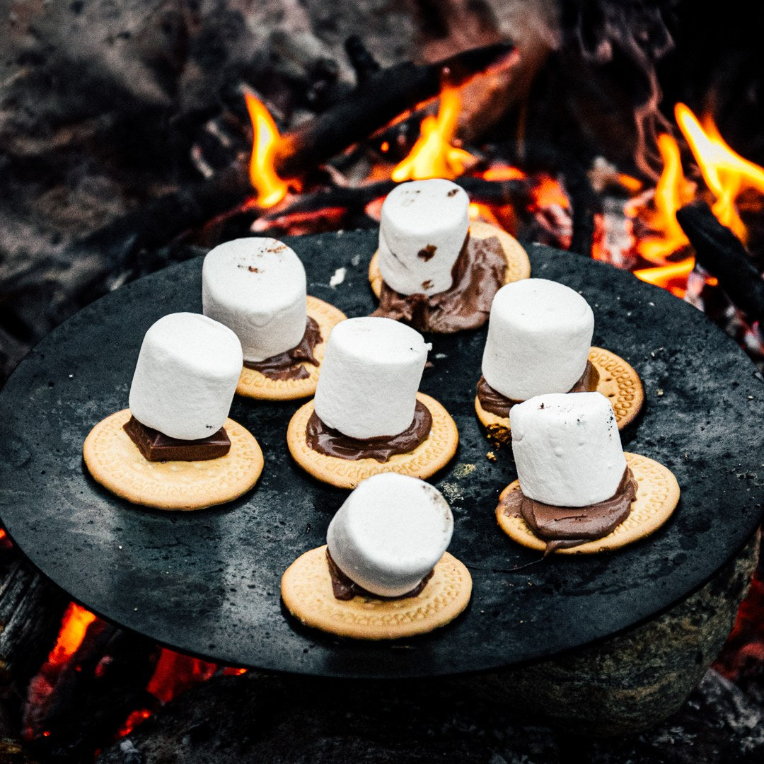 Sju marshmallows på chokladtoppade kex värms på en rund bricka över en öppen lägereld.