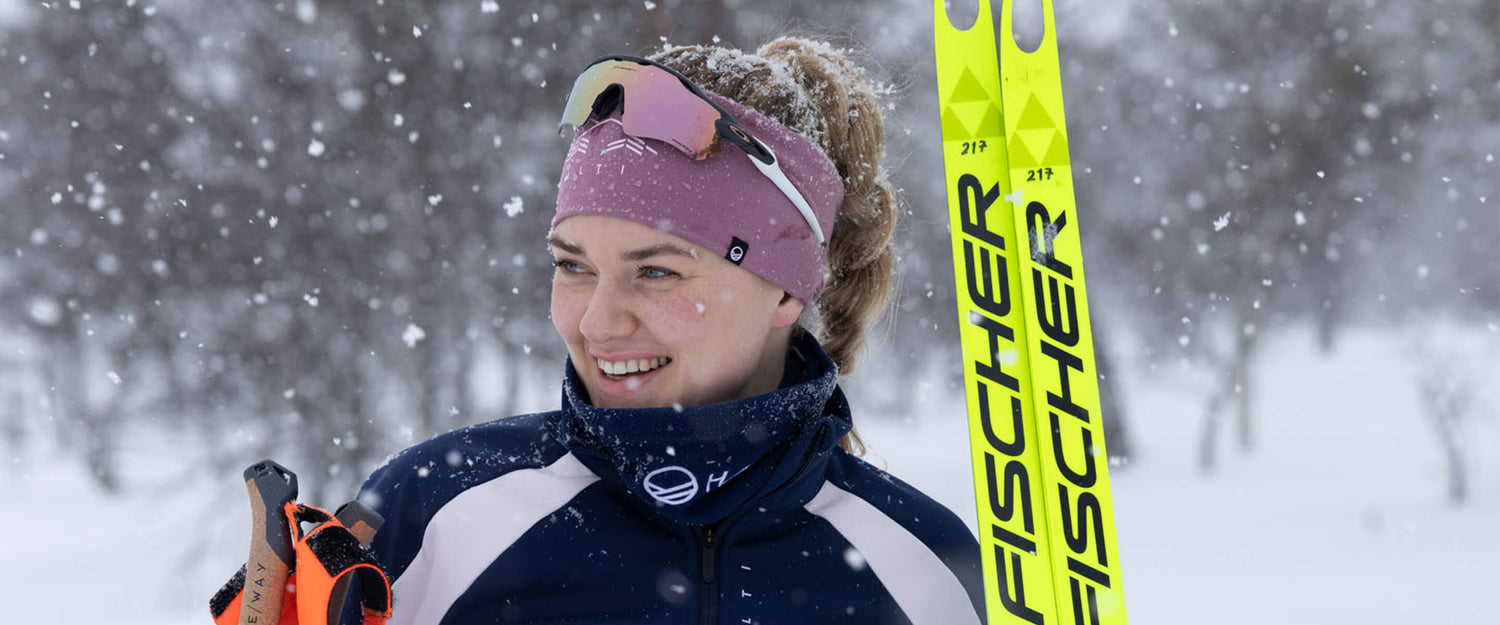 En person i vintersportkläder ler medan han håller i Fischer-skidor i en snöig utomhusmiljö.