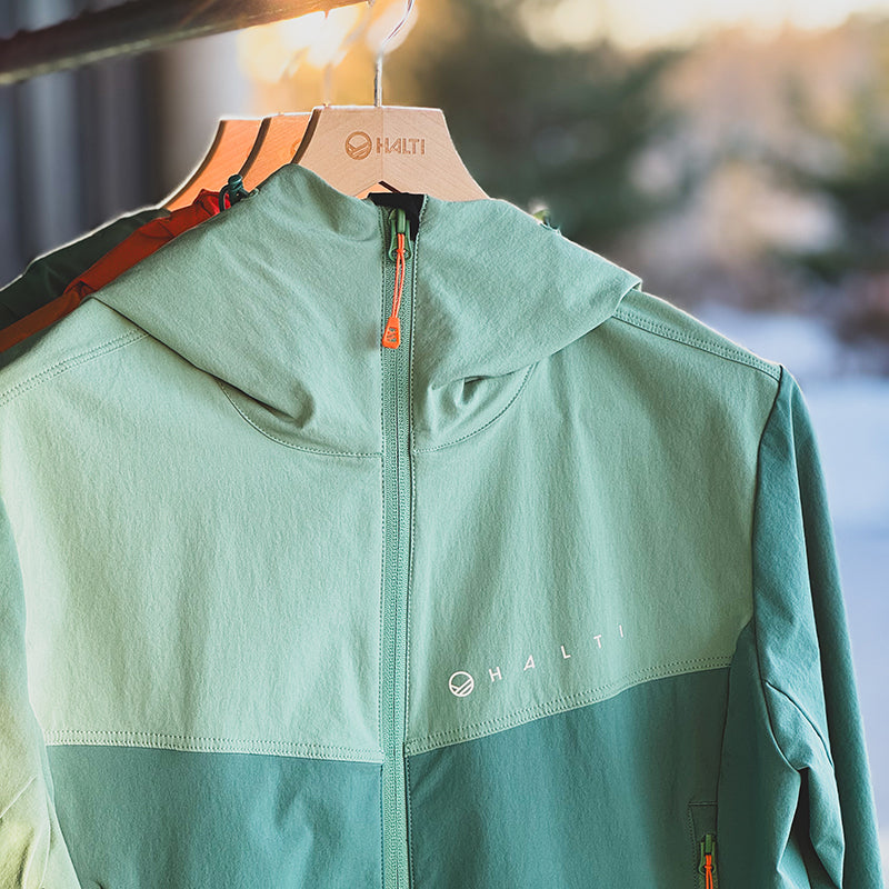 En ljusgrön Halti Pallas Evo X-stretch Jacka Plus Dam hänger utomhus på en trägalge, en annan jacka delvis bakom.