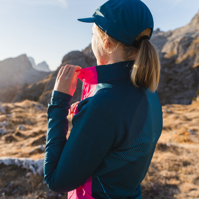 En person i friluftskläder justerar jackans krage medan han står på en klippa med berg i bakgrunden.