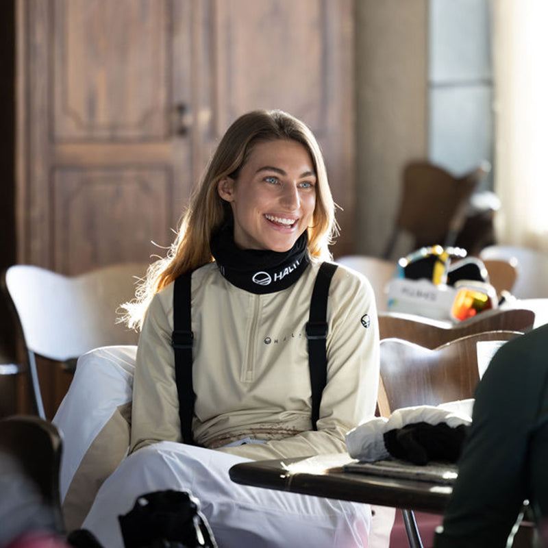 En kvinna i ljus topp och svarta hängslen sitter vid ett bord och ler, med skidutrustning synlig i närheten.