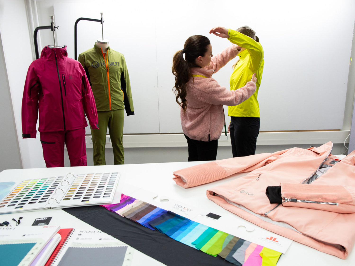 Två personer passar in en gul jacka på en person i en designstudio, med tygprover och jackor på ett bord och skyltdockor i närheten.