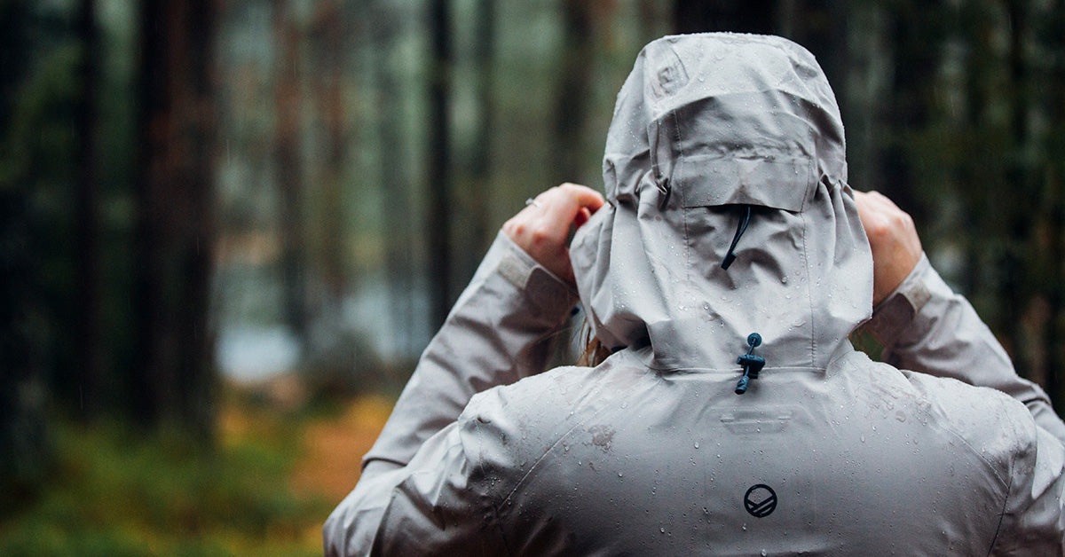 Person i grå regnjacka med huvan uppdragen, står i en regnig skog med suddiga träd i bakgrunden.