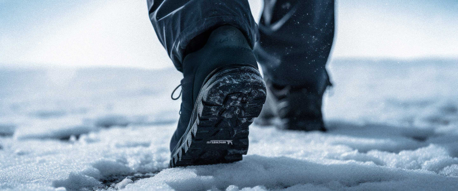 Närbild av en person som går på snö, klädd i mörka byxor och svarta vandringsskor med synligt mönster.