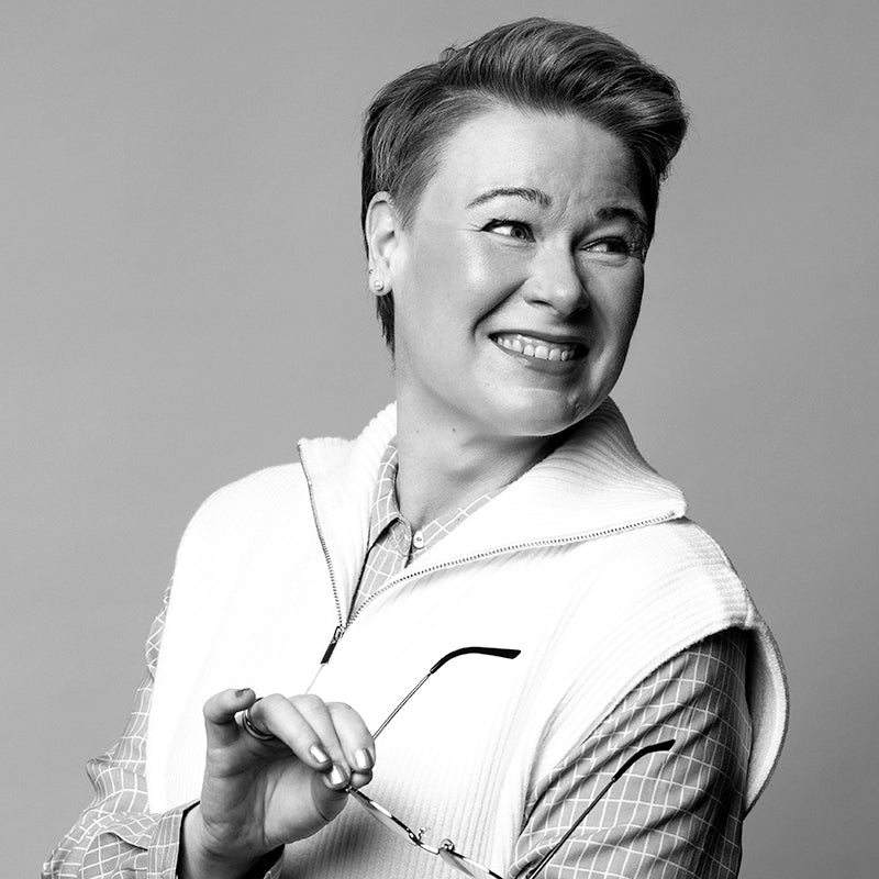 Svartvitt foto av en person som ler, håller i glasögon och bär en väst med dragkedja över en rutig skjorta.