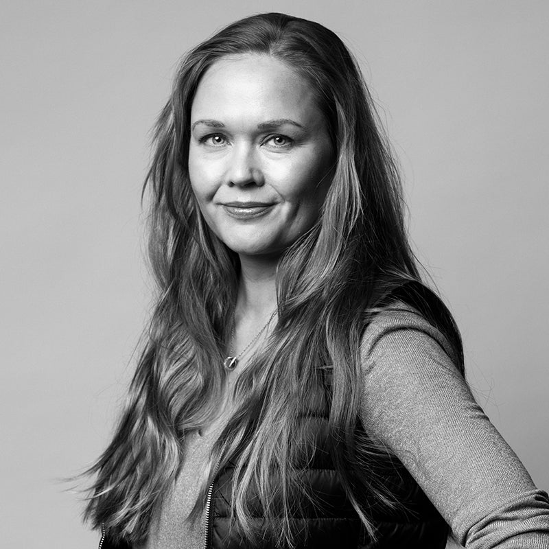 Kvinna med långt hår, klädd i väst och långärmad skjorta, tittar in i kameran mot en enfärgad bakgrund.